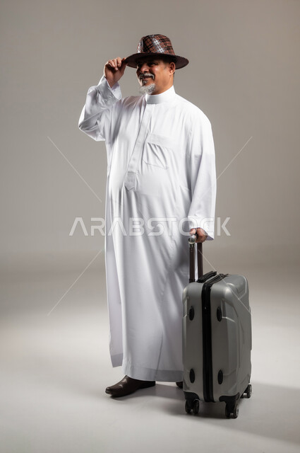 النظر إلى الكاميرا بإيماءات الفرح و السرور , قضاء أوقات ممتعة في الإجازة , الاستمتاع بالسفر و الرحلات الصيفية , بورتريه لرجل عربي خليجي سعودي كبير في السن يرتدي الثوب التقليدي مع قبعة يمسك بيده حقيبة سفر رمادية , صورة للجسم بالطول الكامل , خلفية رمادية