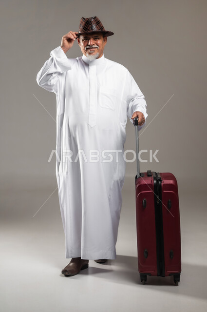 Spending enjoyable times on vacation, enjoying travel and summer trips, portrait of an old Saudi Gulf Arab man wearing a traditional dress with a hat, holding a red suitcase in his hand, looking at the camera with gestures of joy and pleasure, full-length body portrait, gray background