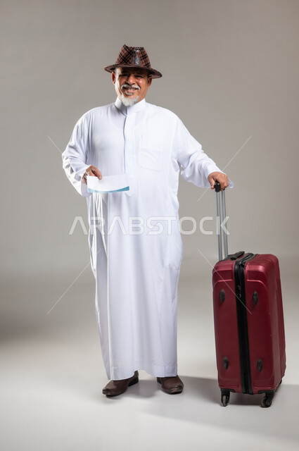 الذهاب في عطلة صيفية ممتعة , التجهيز للسفر و الرحلات , بورتريه لرجل عربي خليجي سعودي كبير في السن يرتدي الثوب التقليدي مع قبعة يمسك بيده حقيبة حمراء و تأشيرة السفر , إيماءات الفرح و السرور , صورة للجسم بالطول الكامل , خلفية رمادية