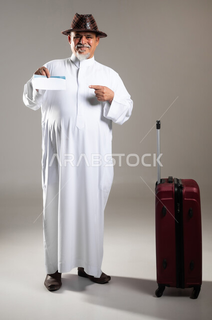 الاستمتاع بالإجازة الصيفية , التجهيز للسفرات و الرحلات الممتعة , بورتريه لرجل عربي خليجي سعودي كبير في السن يرتدي الثوب التقليدي مع قبعة بجانبه حقيبة حمراء يمسك بيده تأشيرة السفر , إيماءات الفرح و الحماس  , صورة للجسم بالطول الكامل , خلفية رمادية