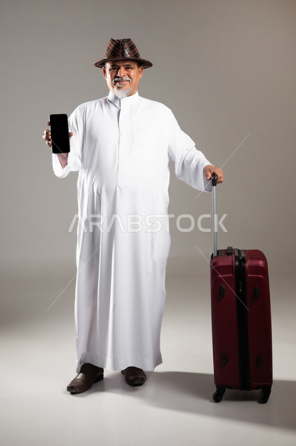 استخدام الأجهزة الحديثة و المتطورة , التجهيز للسفر و الرحلات الممتعة , بورتريه لرجل عربي خليجي سعودي كبير في السن يرتدي الثوب التقليدي مع قبعة يمسك بيده حقيبة سفر حمراء و جهاز الجوال بشاشة سوداء فارغة , صورة للجسم بالطول الكامل , خلفية رمادية