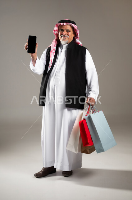 استخدام التقنيات الحديثة للتسوق الالكتروني , شراء الأغراض و الهدايا , بورتريه لرجل عربي خليجي سعودي كبير في السن يرتدي الثوب التقليدي و الشماغ يحمل بيديه أكياس ورقية و جهاز الجوال بشاشة سوداء فارغة , صورة للجسم بالطول الكامل , خلفية رمادية