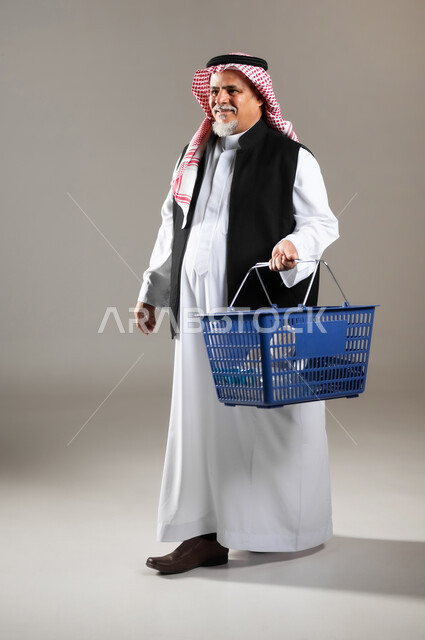 Going to the mall, buying household items and supplies, shopping and getting offers, portrait of an old Saudi Gulf Arab man wearing the traditional thobe and shemagh, holding a shopping basket in his hand, full-length body photo, gray background