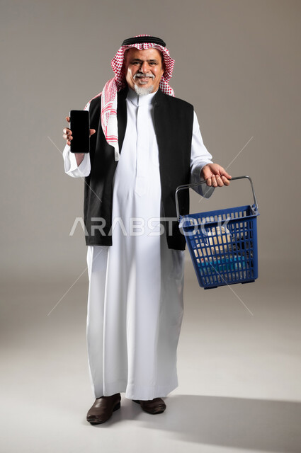 شراء المستلزمات و الأغراض المنزلية , مفهوم التسوق الالكتروني , بورتريه لرجل عربي خليجي سعودي كبير في السن يرتدي الثوب التقليدي و الشماغ يحمل بيده سلة مشتريات و جهاز الجوال بشاشة سوداء فارغة , صورة للجسم بالطول الكامل , خلفية رمادية