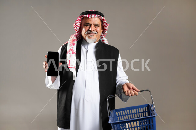 مفهوم التسوق الالكتروني , شراء المستلزمات و الأغراض المنزلية , بورتريه مقرب لرجل عربي خليجي سعودي كبير في السن يرتدي الثوب التقليدي و الشماغ يحمل بيده سلة مشتريات و الهاتف المحمول , عرض شاشة سوداء فارغة عبر الجوال , خلفية رمادية