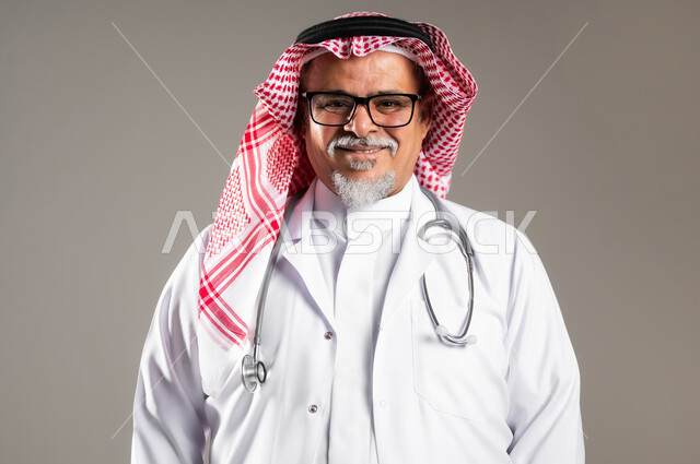العمل في القطاع الطبي , تقديم الخدمات العلاجية , مفهوم الرعاية الصحية , بورتريه مقرب لطبيب عربي خليجي سعودي كبير في السن يرتدي الثوب التقليدي و الشماغ مع المعطف الطبي , النظر إلى الكاميرا بإيماءات الفرح و لسرور , خلفية رمادية