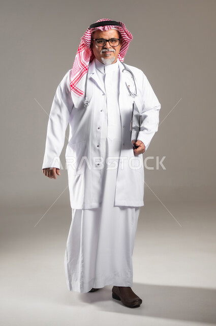 تقديم الخدمات العلاجية للمرضى , العمل في القطاع الصحي , مفهوم الطب والرعاية الصحية , بورتريه لطبيب عربي خليجي سعودي كبير في السن يرتدي الثوب التقليدي و الشماغ مع المعطف الطبي , صورة للجسم بالطول الكامل , خلفية رمادية