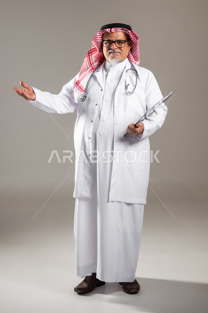 مفهوم الرعاية الصحية , تقديم الخدمات والاحتياجات الخاصة بكل كفاءة , بورتريه لطبيب عربي خليجي سعودي كبير في السن يرتدي الثوب التقليدي و الشماغ مع المعطف الطبي يشير بيده إلى شيء ما , صورة للجسم بالطول الكامل , خلفية رمادية