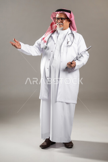 تقديم الخدمات والاحتياجات الخاصة بكل كفاءة , مفهوم الرعاية الصحية , بورتريه لطبيب عربي خليجي سعودي كبير في السن يرتدي الثوب التقليدي و الشماغ مع المعطف الطبي يشير بيده إلى شيء ما , صورة للجسم بالطول الكامل , خلفية رمادية