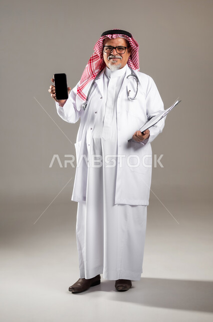 تقديم خدمات الرعاية الصحية , استخدام جهاز الجوال للتواصل مع المرضى , بورتريه لطبيب عربي خليجي سعودي كبير في السن يرتدي الثوب التقليدي و الشماغ مع المعطف الطبي يحمل بيده الهاتف المحمول بشاشة سوداء فارغة , صورة للجسم بالطول الكامل , خلفية رمادية