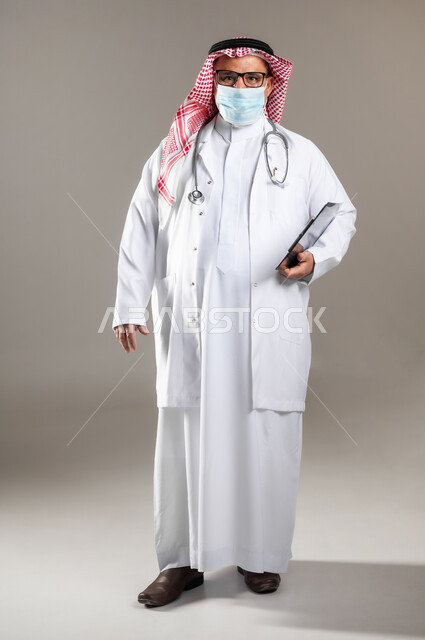 القيام بالفحوصات الطبية اللازمة , تقديم خدمات الرعاية الصحية , بورتريه لطبيب عربي خليجي سعودي كبير في السن يرتدي الثوب التقليدي و الشماغ مع المعطف الطبي و الكمامة يحمل دفتر سجلات المرضى بيده , صورة للجسم بالطول الكامل , خلفية رمادية