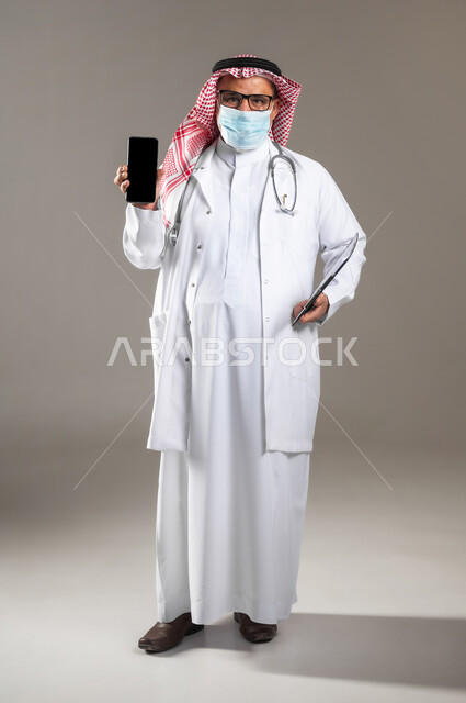 استخدام جهاز الجوال للتواصل مع المرضى , تقديم خدمات الرعاية الصحية , بورتريه لطبيب عربي خليجي سعودي كبير في السن يرتدي الثوب التقليدي و الشماغ مع المعطف الطبي و الكمامة يحمل بيده الهاتف المحمول بشاشة سوداء فارغة , صورة للجسم بالطول الكامل , خلفية رمادية