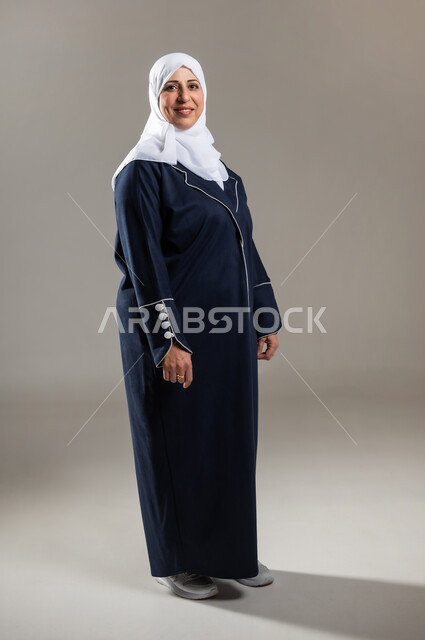 الوقوف باستقامة و استعداد , الصحة البدنية و الجسدية لكبار السن , بورتريه لامرأة عربية خليجية سعودية كبيرة في السن ترتدي الثوب التقليدي و الحجاب , النظر إلى الكاميرا بإيماءات السعادة و السرور , صورة للجسم بالطول الكامل , خلفية رمادية