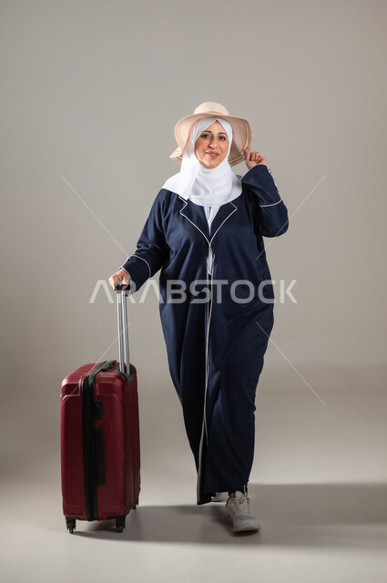 النظر إلى الكاميرا بإيماءات الفرح و السرور , الاستمتاع بالإجازة الصيفية , التجهيز للسفر و الرحلات الممتعة , بورتريه لامرأة عربية خليجية سعودية كبيرة في السن ترتدي الثوب التقليدي و الحجاب مع قبعة من القش تمسك بيدها حقيبة سفر حمراء , صورة للجسم بالطول الكامل , خلفية رمادية