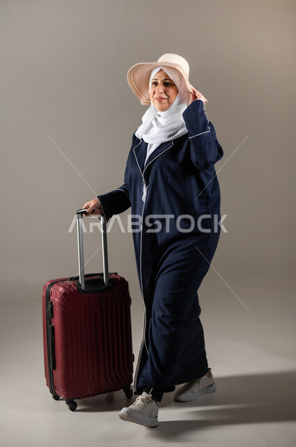 الذهاب في عطلة صيفية ممتعة , التجهيز للسفر و الرحلات , بورتريه لامرأة عربية خليجية سعودية كبيرة في السن ترتدي الثوب التقليدي و الحجاب مع قبعة من القش تمسك بيدها حقيبة سفر حمراء , النظر بإيماءات الفرح و السرور , صورة للجسم بالطول الكامل , خلفية رمادية