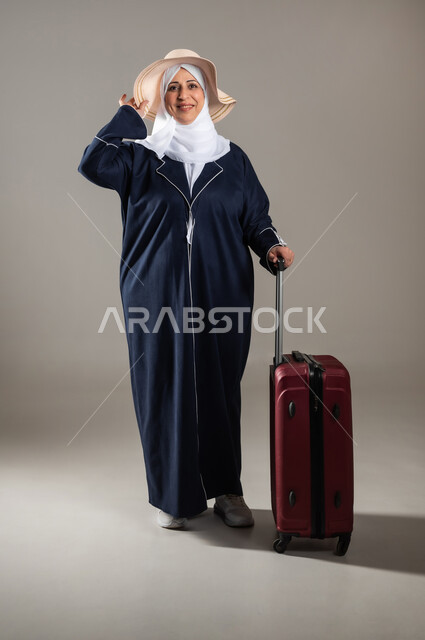 الاستمتاع بالإجازة الصيفية , التجهيز للسفر و الرحلات الممتعة , بورتريه لامرأة عربية خليجية سعودية كبيرة في السن ترتدي الثوب التقليدي و الحجاب مع قبعة من القش تمسك بيدها حقيبة سفر حمراء , النظر بإيماءات الفرح و السرور , صورة للجسم بالطول الكامل , خلفية رمادية