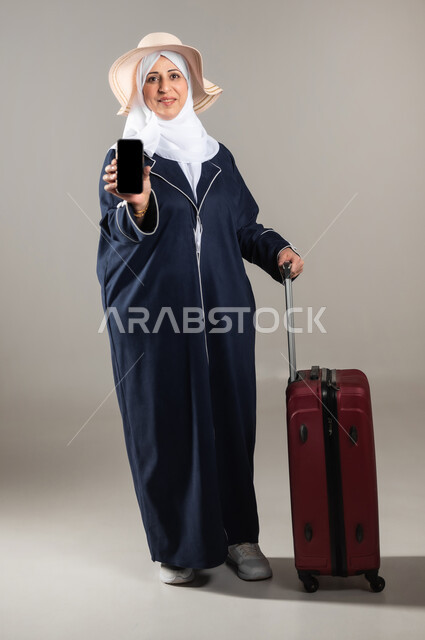 استخدام الأجهزة الحديثة و المتطورة , التجهيز للسفر و الرحلات الممتعة , بورتريه لامرأة عربية خليجية سعودية كبيرة في السن ترتدي الثوب التقليدي و الحجاب مع قبعة من القش تمسك بيديها حقيبة سفر حمراء و الهاتف المحمول بشاشة سوداء فارغة , صورة للجسم بالطول الكامل , خلفية رمادية