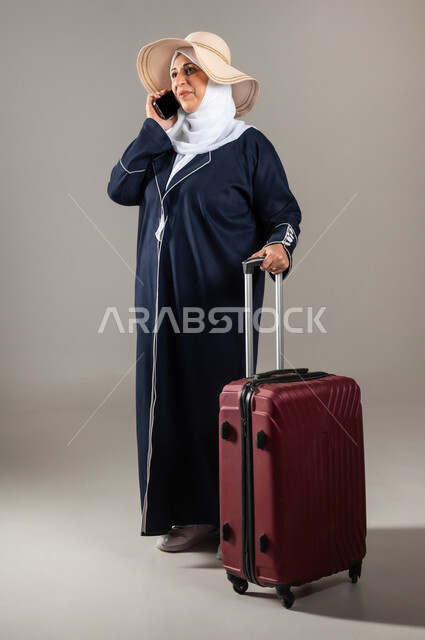 Preparing for travel and enjoyable trips, making a phone call via mobile phone, sharing good news with family, portrait of an elderly Saudi Gulf Arab woman wearing traditional dress and hijab, holding a red suitcase in her hand, full-length body portrait, gray background
