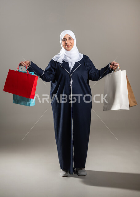 النظر بإيماءات الفرح و السرور , شراء الأغراض الجديدة و المستلزمات , الاستمتاع بالتسوق و الحصول على العروض , بورتريه لامرأة عربية خليجية سعودية كبيرة في السن ترتدي الثوب التقليدي و الحجاب تحمل الأكياس الورقية بيديها , صورة للجسم بالطول الكامل , خلفية رمادية