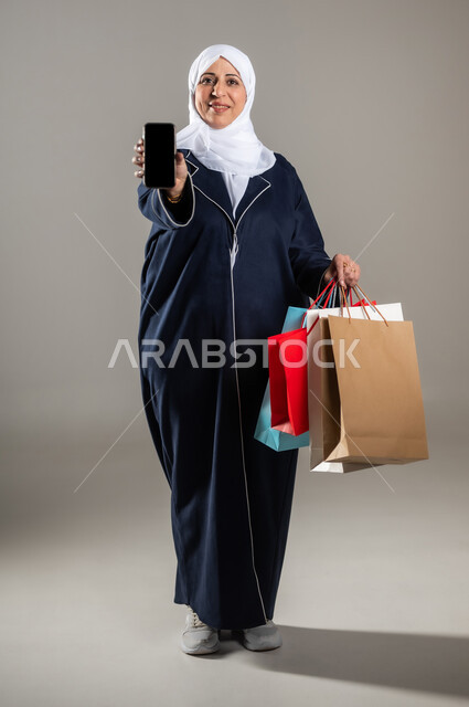 Shopping and purchasing household supplies, using modern technical devices, portrait of an elderly Saudi Arabian Gulf woman wearing a traditional dress and hijab, holding shopping bags and a mobile phone in her hand with a blank black screen, full-length body photo, gray background