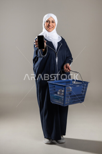 استخدام الأجهزة التقنية الحديثة , التسوق و شراء المستلزمات المنزلية , بورتريه لامرأة عربية خليجية سعودية كبيرة في السن ترتدي الثوب التقليدي و الحجاب تحمل بيدها الهاتف المحمول بشاشة سوداء فارغة , صورة للجسم بالطول الكامل , خلفية رمادية
