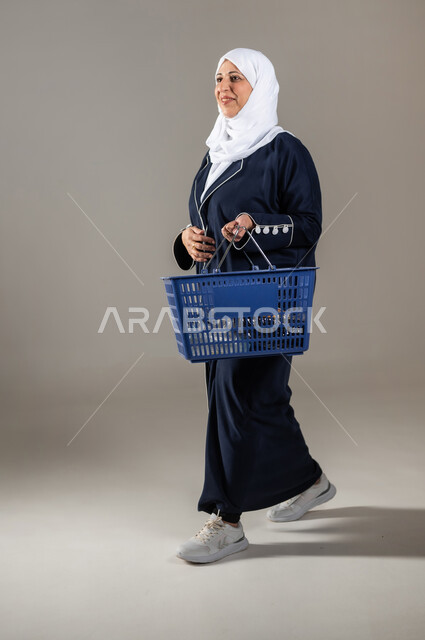 الذهاب إلى المجمع التجاري , شراء الأغراض و المستلزمات المنزلية , التسوق والحصول على العروض , بورتريه لامرأة عربية خليجية سعودية كبيرة في السن ترتدي الثوب التقليدي و الحجاب تحمل سلة مشتريات , صورة للجسم بالطول الكامل , خلفية رمادية