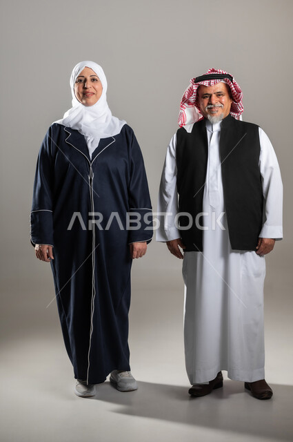 مفهوم التقدم بالعمر , المحافظة على الصحة البدنية لكبار السن , بورتريه لزوجان عربيان خليجيان سعوديان كبيران في السن يرتديان الثوب التقليدي يقفان باستقامة , النظر إلى الكاميرا بإيماءات السعادة و السرور , صورة للجسم بالطول الكامل , خلفية رمادية