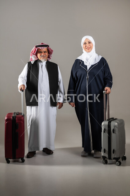 مفهوم السياحة والقيام بالجولات الترفيهية وقت الإجازة, الوقوف باستقامة وانتظار موعد الطائرة, بورتريه لزوجان عربيان خليجيان سعوديان كبار بالسن  يرتديان الزي السعودي التقليدي يمسكان بيدهما حقائب السفر للاستعداد لبدء الرحلة في العطلة الصيفيه, صورة الجسم بالطول الكامل, خلفية رماديه