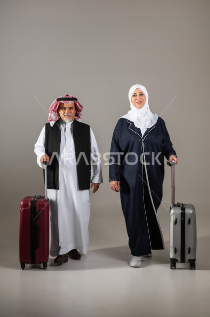 مفهوم السياحة والقيام بالجولات الترفيهية وقت الإجازة, الوقوف باستقامة وانتظار موعد الطائرة, بورتريه لزوجان عربيان خليجيان سعوديان كبار بالسن  يرتديان الزي السعودي التقليدي يمسكان بيدهما حقائب السفر للاستعداد لبدء الرحلة في العطلة الصيفيه, صورة الجسم بالطول الكامل, خلفية رماديه