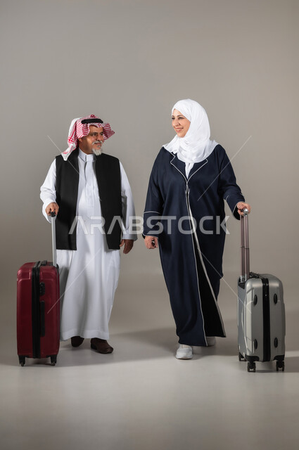مفهوم السياحة والقيام بالجولات الترفيهية وقت الإجازة, المشي والذهاب الى مكان صعود الطائره, بورتريه لزوجان عربيان خليجيان سعوديان كبار بالسن  يرتديان الزي السعودي التقليدي يمسكان بيدهما حقائب السفر للاستعداد لبدء الرحلة في العطلة الصيفيه, صورة الجسم بالطول الكامل, خلفية رماديه