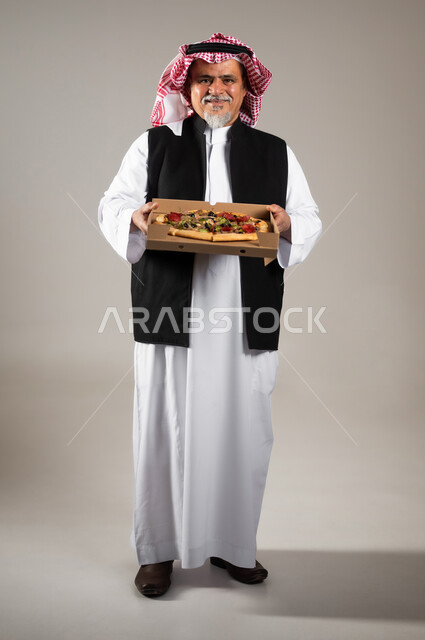 أصناف المعجنات والفطائر الشهية، طلبات الأطعمة الخارجية، بورتريه بالطول الكامل لرجل عربي خليجي سعودي كبير بالسن يرتدي الثوب التقليدي والشماغ والستره الوقائيه يحمل البيتزا في علبة كرتون، أضرار تناول وجبات طعام غير صحية، مطاعم المأكولات الغربية في السعودية، خلفية رمادية