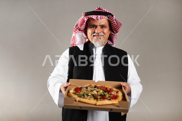 أصناف المعجنات والفطائر الشهية، طلبات الأطعمة الخارجية، بورتريه بالطول مقرب لرجل عربي خليجي سعودي كبير بالسن يرتدي الثوب التقليدي والشماغ والستره الوقائيه يحمل البيتزا في علبة كرتون، أضرار تناول وجبات طعام غير صحية، مطاعم المأكولات الغربية في السعودية، خلفية رمادية