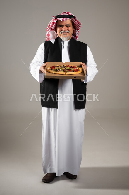 أصناف المعجنات والفطائر الشهية، طلبات الأطعمة الخارجية، بورتريه بالطول الكامل لرجل عربي خليجي سعودي كبير بالسن يرتدي الثوب التقليدي والشماغ والستره الوقائيه يحمل البيتزا في علبة كرتون، أضرار تناول وجبات طعام غير صحية، مطاعم المأكولات الغربية في السعودية، خلفية رمادية