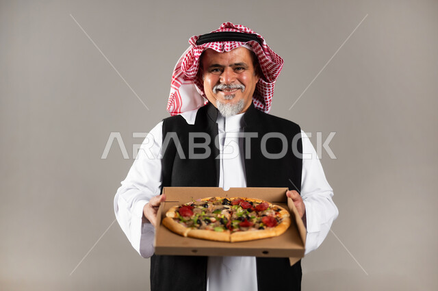 أصناف المعجنات والفطائر الشهية، طلبات الأطعمة الخارجية، بورتريه مقرب لرجل عربي خليجي سعودي كبير بالسن يرتدي الثوب التقليدي والشماغ والستره الوقائيه يحمل البيتزا في علبة كرتون، أضرار تناول وجبات طعام غير صحية، مطاعم المأكولات الغربية في السعودية، خلفية رمادية