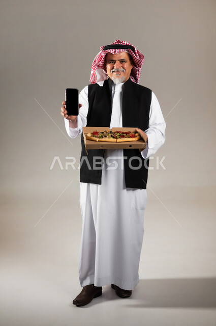 طلبات الأطعمة الخارجية عبر تطبيقات الجوال، بورتريه  لرجل عربي خليجي سعودي كبير بالسن يرتدي الثوب التقليدي والشماغ والستره الوقائيه يحمل البيتزا في علبة كرتون ويشير بهاتفه المحمول بشاشه سوداء فارغه، أضرار تناول وجبات طعام غير صحية، مطاعم المأكولات الغربية في السعودية، خلفية رمادية