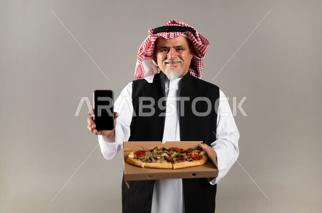 طلبات الأطعمة الخارجية عبر تطبيقات الجوال، بورتريه  لرجل عربي خليجي سعودي كبير بالسن يرتدي الثوب التقليدي والشماغ والستره الوقائيه يحمل البيتزا في علبة كرتون ويشير بهاتفه المحمول بشاشه سوداء فارغه، أضرار تناول وجبات طعام غير صحية، مطاعم المأكولات الغربية في السعودية، خلفية رمادية
