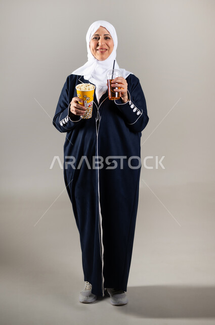 الاستمتاع بتناول الفشار والمشروبات البارده، بورتريه لسيدة عربية خليجية سعودية كبيره بالسن ترتدي الحجاب والعباءه السوداء تمسك بيدها المشروب الغازي و الفشار ، ايماءات الوجه واليدين تدل على الاستمتاع والحماس، سناكات ومقرمشات، مشروبات غازيه و عصائر، خلفيه رماديه