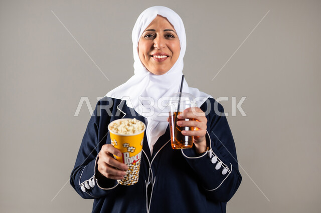 الاستمتاع بتناول الفشار والمشروبات البارده، بورتريه مقرب لسيدة عربية خليجية سعودية كبيره بالسن ترتدي الحجاب والعباءه السوداء تمسك بيدها المشروب الغازي و الفشار ، ايماءات الوجه واليدين تدل على الاستمتاع والحماس، سناكات ومقرمشات، مشروبات غازيه و عصائر، خلفيه رماديه