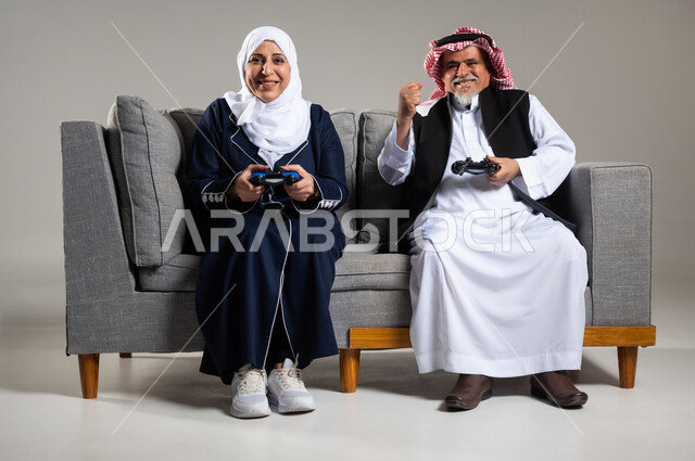 The concept of spending happy times playing with family, portrait of an elderly Saudi Arabian Gulf couple sitting on a comfortable sofa in the living room holding a joystick and playing video games, facial gestures indicating challenge, fun and entertainment with video games, PlayStation entertainment games, group play, entertainment and recreation, technology and games, gray background