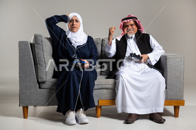 مفهوم قضاء الاوقات السعيده باللعب مع الاهل، بورتريه لزوجان عربيان سعوديان خليجيان كبيران بالسن يجلسان على كنبه مريحه في غرفه المعيشه يمسكان عصا التحكم و يلعبان بألعاب الفيديو، إيماءات وجه تدل على التحدي، المتعة و التسلية بألعاب الفيديو، ألعاب البلايستيشن الترفيهية، اللعب الجماعي, تسليه وترفيه, تقنيات و ألعاب، خلفيه رماديه