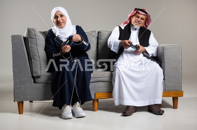 مفهوم قضاء الاوقات السعيده باللعب مع الاهل، بورتريه لزوجان عربيان سعوديان خليجيان كبيران بالسن يجلسان على كنبه مريحه في غرفه المعيشه يمسكان عصا التحكم و يلعبان بألعاب الفيديو، إيماءات وجه تدل على التحدي، المتعة و التسلية بألعاب الفيديو، ألعاب البلايستيشن الترفيهية، اللعب الجماعي, تسليه وترفيه, تقنيات و ألعاب، خلفيه رماديه