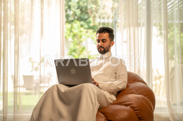 استعمال جهاز الحاسوب المحمول للعمل عن بعد، التطور التقني والتكنولوجي في مختلف المجالات، إنجاز الأعمال بسرعة وسهولة، عقد الاجتماعات عن طريق الانترنت، رجل عربي خليجي سعودي يجلس على أريكة في غرفة المعيشة ويقوم بتصفح اللابتوب