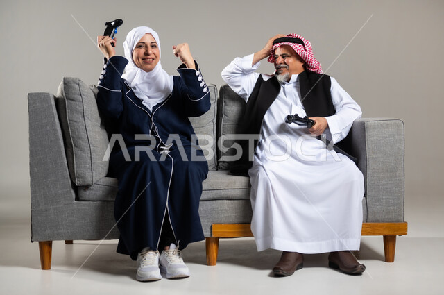 مفهوم قضاء الاوقات السعيده باللعب مع الاهل، بورتريه لزوجان عربيان سعوديان خليجيان كبيران بالسن يجلسان على كنبه مريحه في غرفه المعيشه يمسكان عصا التحكم و يلعبان بألعاب الفيديو، إيماءات وجه تدل على التحدي، المتعة و التسلية بألعاب الفيديو، ألعاب البلايستيشن الترفيهية، اللعب الجماعي, تسليه وترفيه, تقنيات و ألعاب، خلفيه رماديه