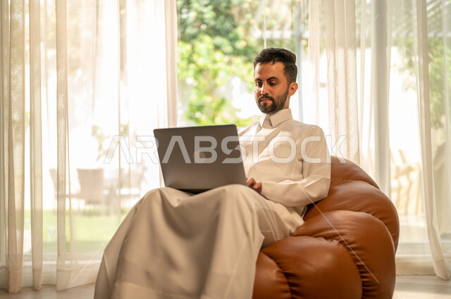 التطور التقني والتكنولوجي في مختلف المجالات، استخدام جهاز الحاسوب المحمول للعمل عن بعد، إنجاز الأعمال بسرعة وسهولة، عقد الاجتماعات عن طريق الانترنت، رجل عربي خليجي سعودي يجلس على أريكة في غرفة المعيشة ويقوم بتصفح اللابتوب