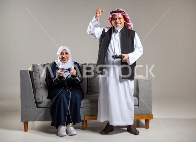 The concept of spending happy times playing with family, portrait of an elderly Saudi Arabian Gulf couple sitting on a comfortable sofa in the living room holding a joystick and playing video games, facial gestures indicating challenge, fun and entertainment with video games, PlayStation entertainment games, group play, entertainment and recreation, technology and games, gray background