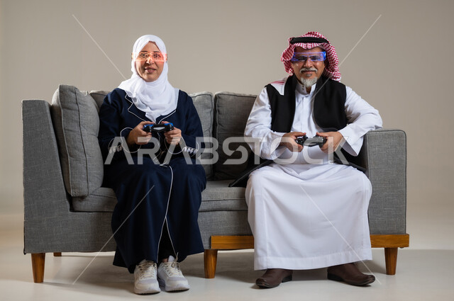 مفهوم قضاء الاوقات السعيده باللعب مع الاهل، بورتريه لزوجان عربيان سعوديان خليجيان كبيران بالسن يجلسان على كنبه مريحه في غرفه المعيشه يمسكان عصا التحكم و يلعبان بألعاب الفيديو، إيماءات وجه تدل على التحدي، المتعة و التسلية بألعاب الفيديو، ألعاب البلايستيشن الترفيهية، اللعب الجماعي, تسليه وترفيه, تقنيات و ألعاب، خلفيه رماديه