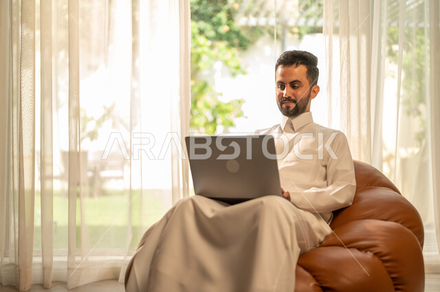 إنجاز الأعمال بسرعة وسهولة، عقد الاجتماعات عن طريق الانترنت، استخدام جهاز الحاسوب المحمول للعمل عن بعد، التطور التقني والتكنولوجي في مختلف المجالات، رجل عربي خليجي سعودي يجلس على أريكة في غرفة المعيشة ويقوم بتصفح اللابتوب