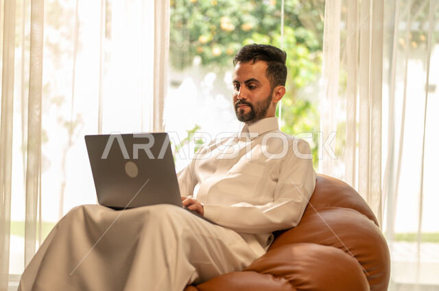 عقد الاجتماعات عن طريق الانترنت، إنجاز الأعمال بسرعة وسهولة، استخدام جهاز الحاسوب المحمول للعمل عن بعد، التطور التقني والتكنولوجي في مختلف المجالات، رجل عربي خليجي سعودي يجلس على أريكة في غرفة المعيشة ويقوم بتصفح اللابتوب
