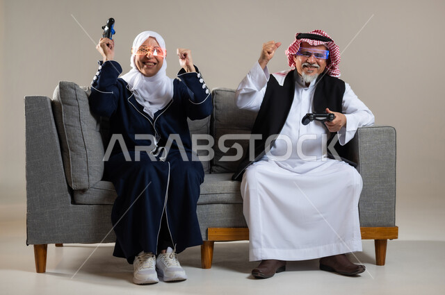 مفهوم قضاء الاوقات السعيده باللعب مع الاهل، بورتريه لزوجان عربيان سعوديان خليجيان كبيران بالسن يجلسان على كنبه مريحه في غرفه المعيشه يمسكان عصا التحكم و يلعبان بألعاب الفيديو، إيماءات وجه تدل على التحدي، المتعة و التسلية بألعاب الفيديو، ألعاب البلايستيشن الترفيهية، اللعب الجماعي, تسليه وترفيه, تقنيات و ألعاب، خلفيه رماديه