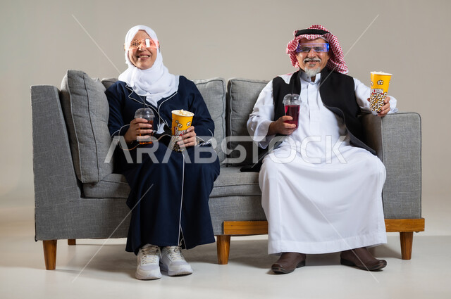 تعابير الحماس والسعادة، التسليه بالسناكس اثناء مشاهده التلفاز، بورتريه لزوجان عربيان خليجيان سعوديان كبار بالسن يجلسان على كنبة مريحة يقومان بمتابعة التلفازوتناول الفشار والعصائر المثلجه، الاستمتاع بقضاء أوقات ترفيهيه، أنشطة عطلة نهاية الأسبوع، خلفية رماديه